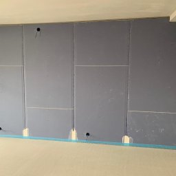 Remonty Wykonczenia - Wnętrze w trakcie prac: ściana pokryta szarymi płytami, widoczne otwory na instalacje elektryczne, zabezpieczona podłoga taśmą malarską.