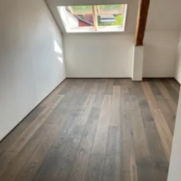 Poddasze z nowo ułożonymi panelami podłogowymi w odcieniach brązu i szarości, widoczne okno dachowe i drewniana belka konstrukcyjna.