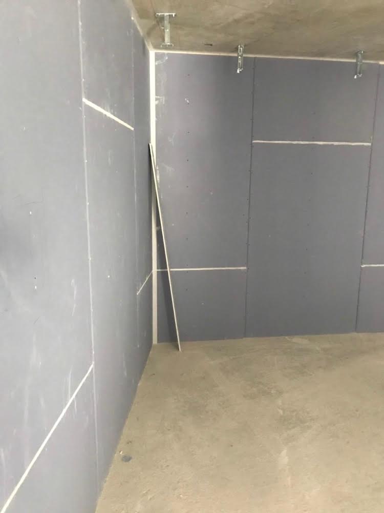 Wnętrze w surowym stanie: ściany wyłożone płytami, widoczne łączenia, surowy betonowy sufit z metalowymi profilami montażowymi.