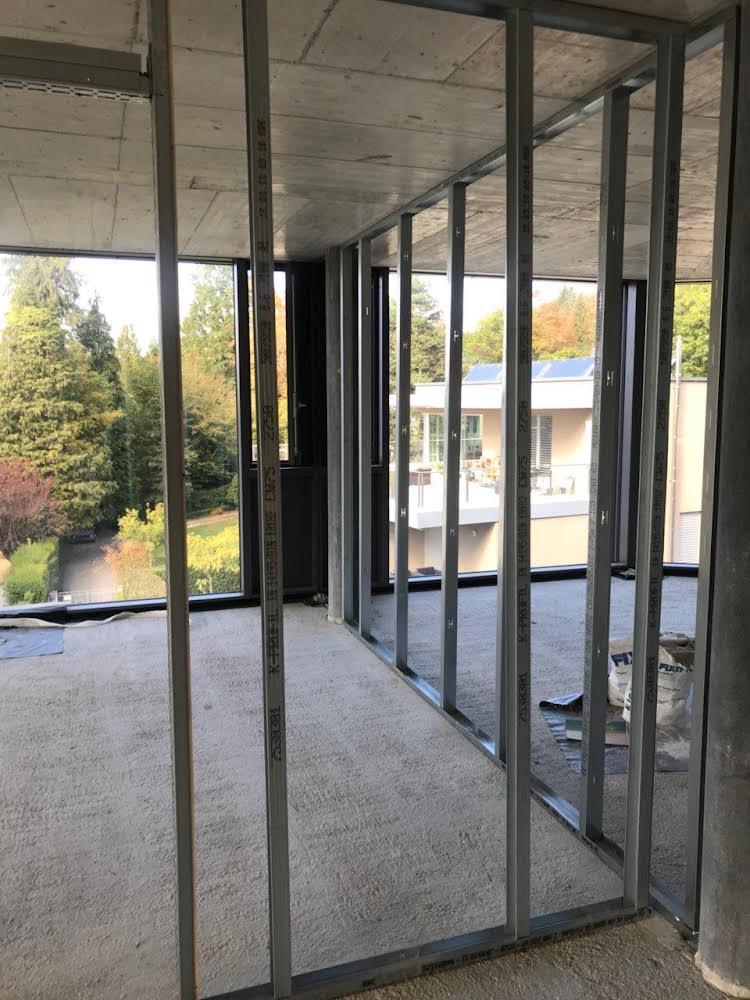 Montaż metalowej konstrukcji ścianki działowej z płyty karton gips w surowym wnętrzu z widokiem na zieleń za oknem.