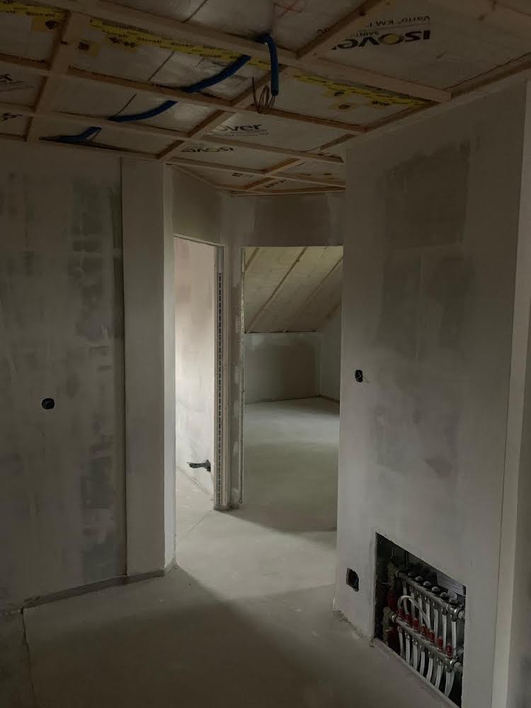 Wnętrze w trakcie prac wykończeniowych: surowa ściana z instalacją ogrzewania podłogowego, widoczne przewody na suficie podwieszanym z izolacją, otwarte przejścia do kolejnych pomieszczeń ze...