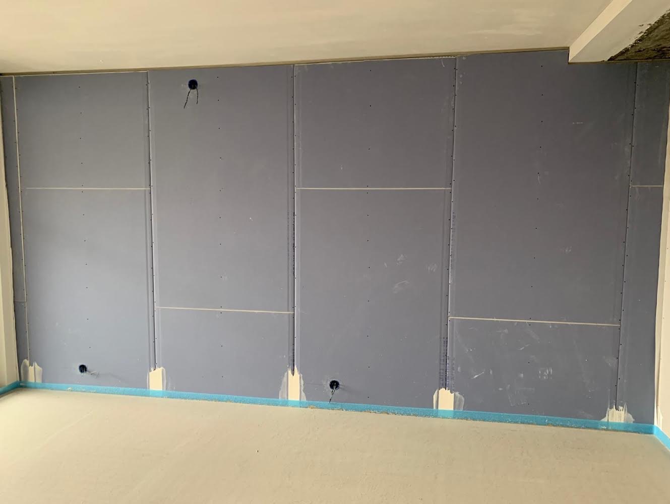 Wnętrze w trakcie prac: ściana pokryta szarymi płytami, widoczne otwory na instalacje elektryczne, zabezpieczona podłoga taśmą malarską.