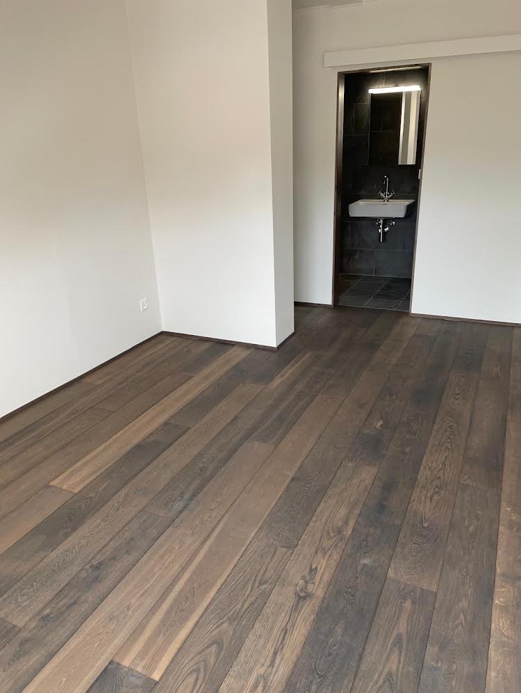 Ułożone panele podłogowe z ciemnego drewna w jasnym pomieszczeniu z widokiem na łazienkę z umywalką i czarnymi płytkami.