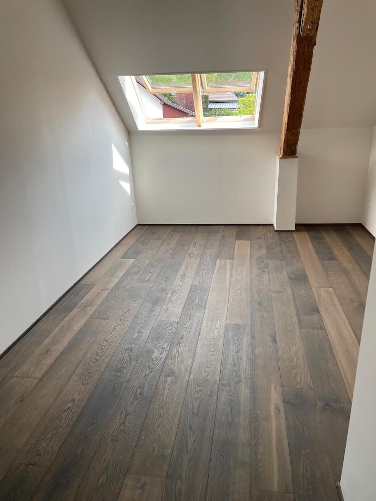 Poddasze z nowo ułożonymi panelami podłogowymi w odcieniach brązu i szarości, widoczne okno dachowe i drewniana belka konstrukcyjna.