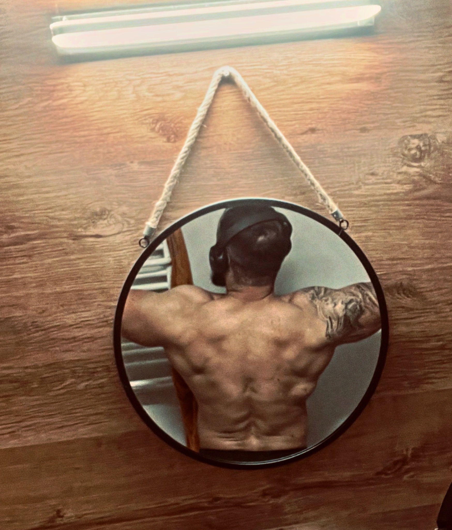 Mężczyzna z tatuażem na ramieniu, widoczny w okrągłym lustrze wiszącym na drewnianej ścianie, prezentuje umięśnione plecy.