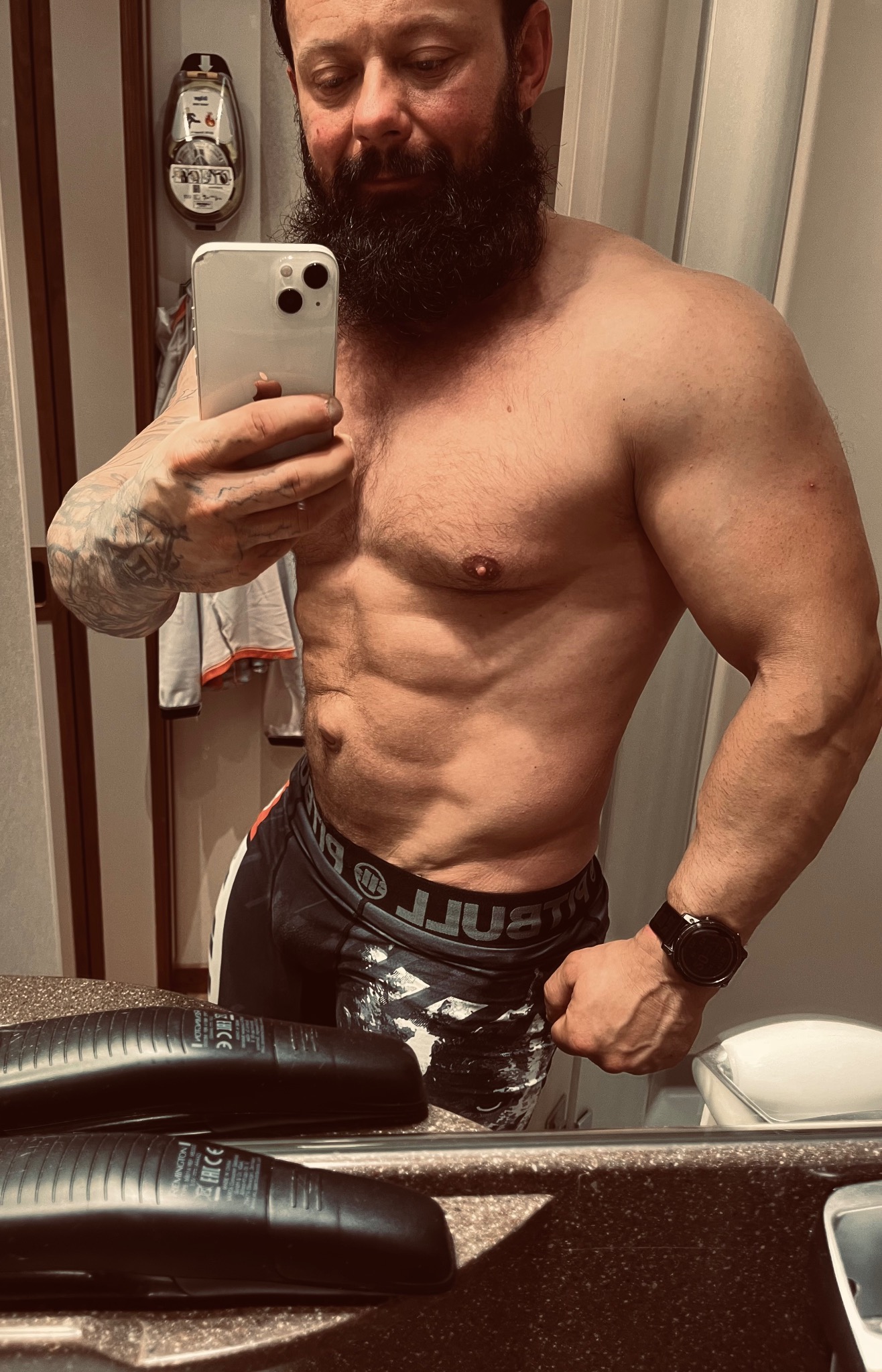 Mężczyzna z brodą robi selfie, prezentując wyrzeźbioną sylwetkę i tatuaż na ramieniu w łazience z widocznymi kosmetykami na blacie.