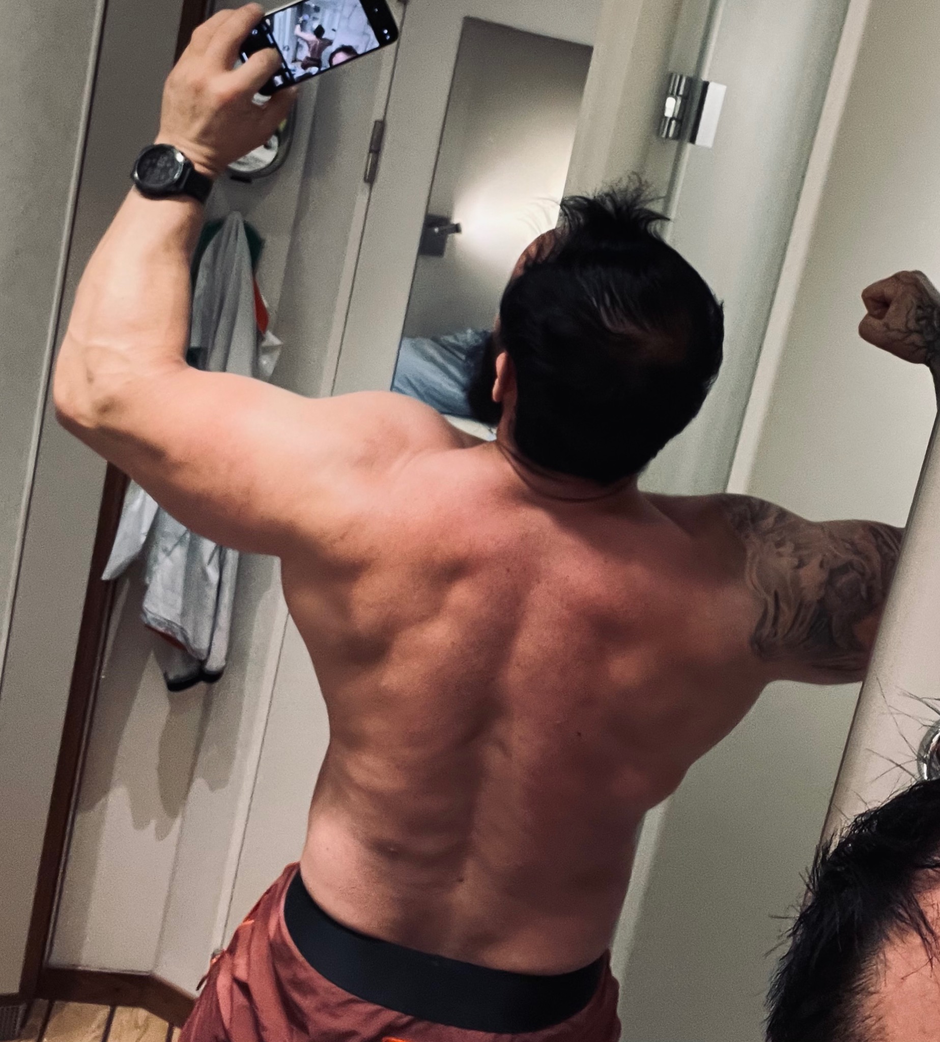 Mężczyzna z widocznymi mięśniami pleców i ramion, robiący sobie zdjęcie telefonem w lustrze. Widoczny tatuaż na ramieniu.