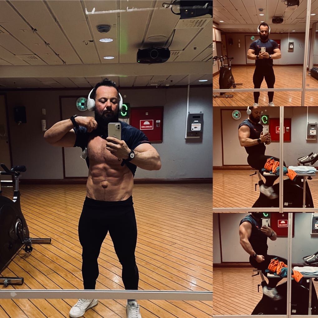 Mężczyzna z brodą w słuchawkach, robi selfie w lustrze, podnosząc koszulkę, aby pokazać wyrzeźbiony brzuch w siłowni z drewnianą podłogą i sprzętem do ćwiczeń.