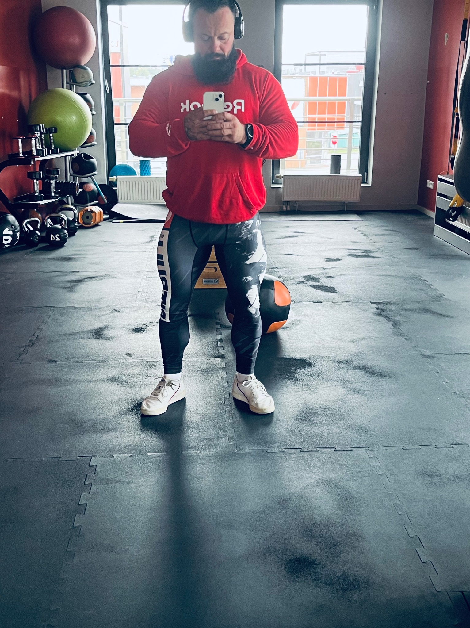 Mężczyzna z brodą w czerwonej bluzie Reebok i słuchawkach, przegląda telefon w siłowni, w tle sprzęt do ćwiczeń i okno.