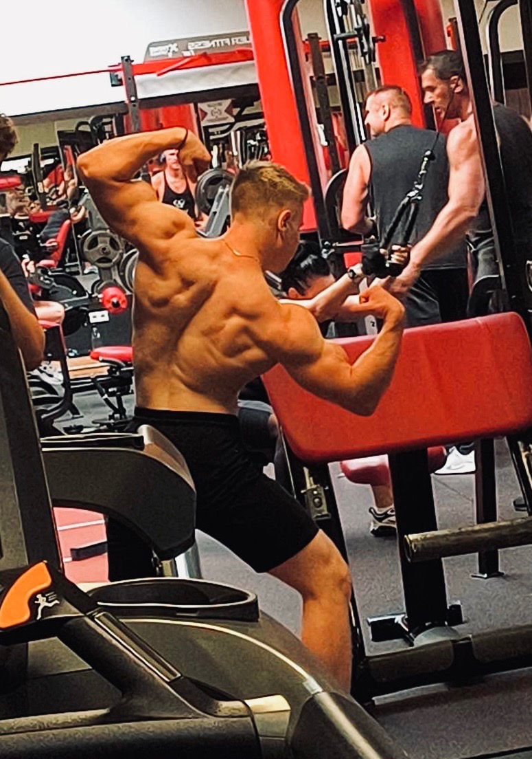 Mężczyzna z odsłoniętymi plecami, prezentujący muskulaturę w siłowni, z widocznym sprzętem do ćwiczeń i innymi osobami w tle.