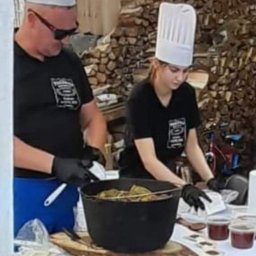 Catering Pracownia Wędzarnicza - Dwoje kucharzy w czarnych koszulkach i białych czapkach przygotowuje potrawy na stole z białym obrusem. Na stole znajdują się deski z przekąskami, duży garnek z jedzeniem i małe pojemniki z sosem.