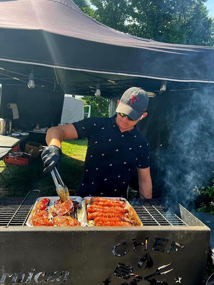 Kucharz w czarnej koszulce z wzorem i czapce bejsbolowej, w okularach przeciwsłonecznych, grilluje mięso i kiełbaski na zewnątrz pod czarnym namiotem; dym unosi się znad grilla z napisem 'Chef'.