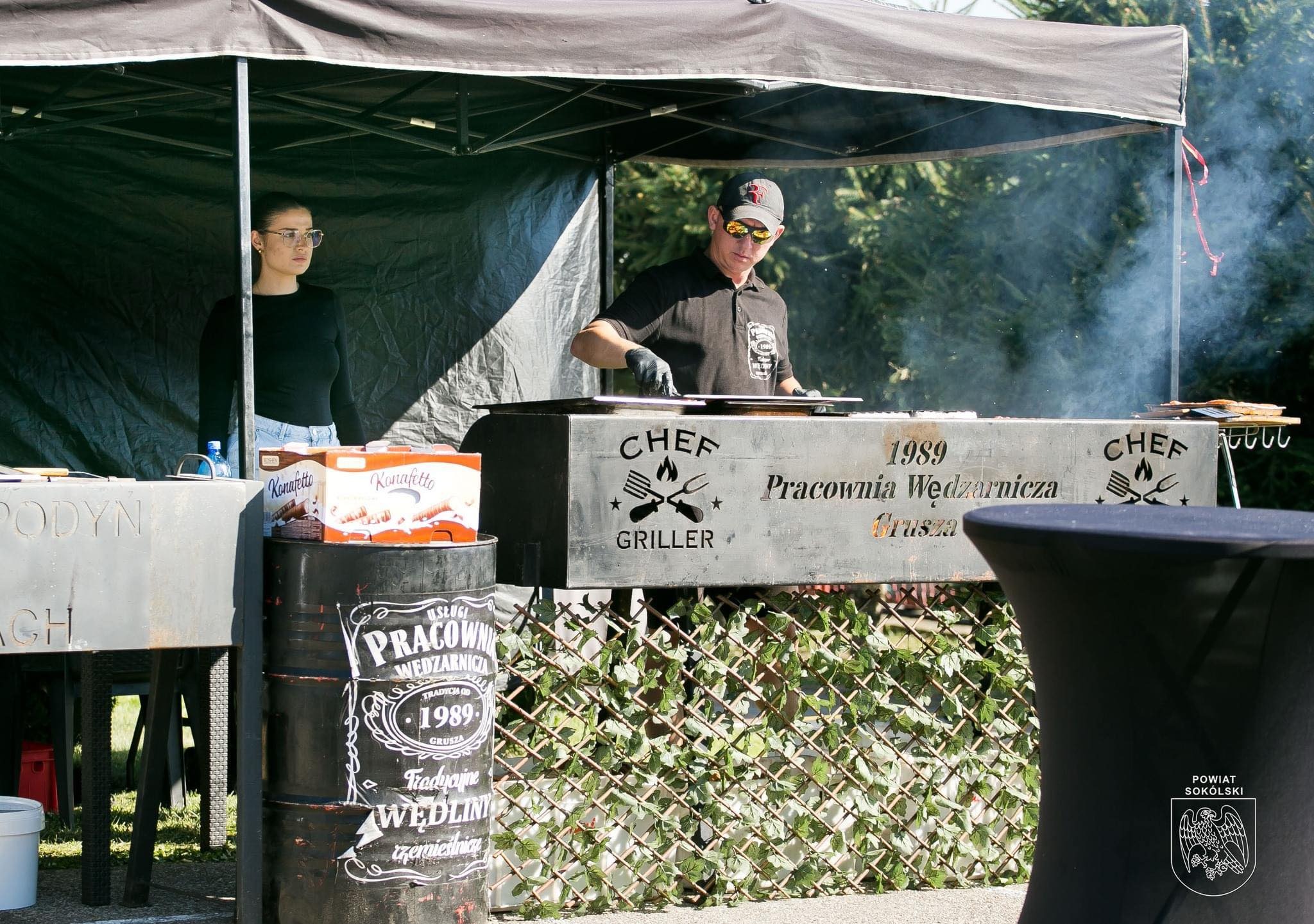 Przygotowywanie wędlin na stoisku cateringowym z napisem 'Pracownia Wędzarnicza Grusza 1989', widoczna osoba w czapce i okularach przeciwsłonecznych w trakcie obsługi grilla, obok skrzynki...