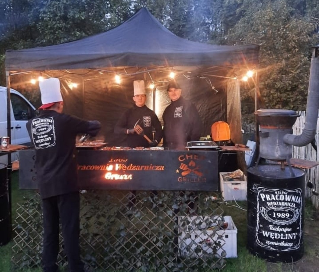 Mobilne stoisko gastronomiczne z grillem i wędzarnią, obsługa w czapkach kucharskich, logo firmy na stoisku i wędzarni, wieczorne oświetlenie żarówkami.