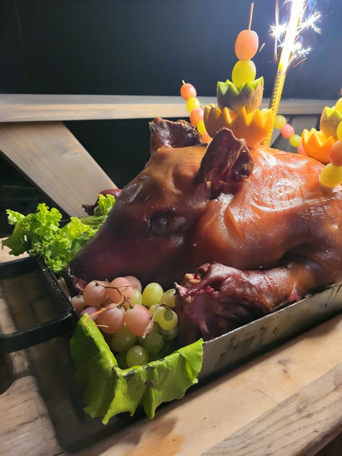 Pieczone prosię udekorowane owocami i racą, podane na liściach sałaty i z winogronami, na drewnianym stole.