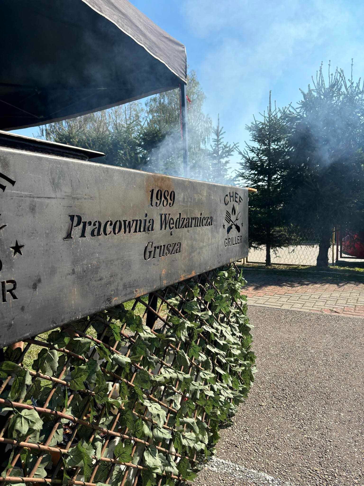 Metalowa konstrukcja wędzarni z napisem 'Pracownia Wędzarnicza Grusza 1989 Chef Griller', ozdobiona sztucznym bluszczem, z widocznym dymem i drzewami w tle.