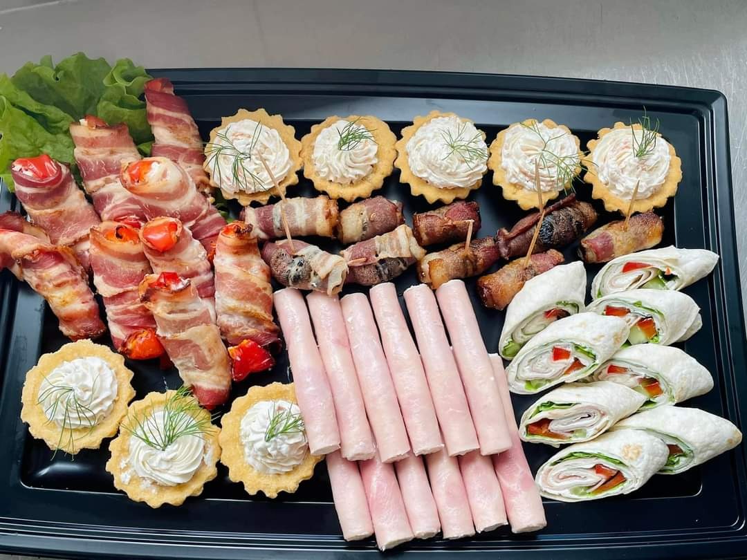 Czarna taca z różnorodnymi przekąskami: roladki z boczkiem, tartaletki z kremowym nadzieniem i koperkiem, mini-szaszłyki owinięte boczkiem, roladki z tortilli z warzywami i szynką oraz szynka...