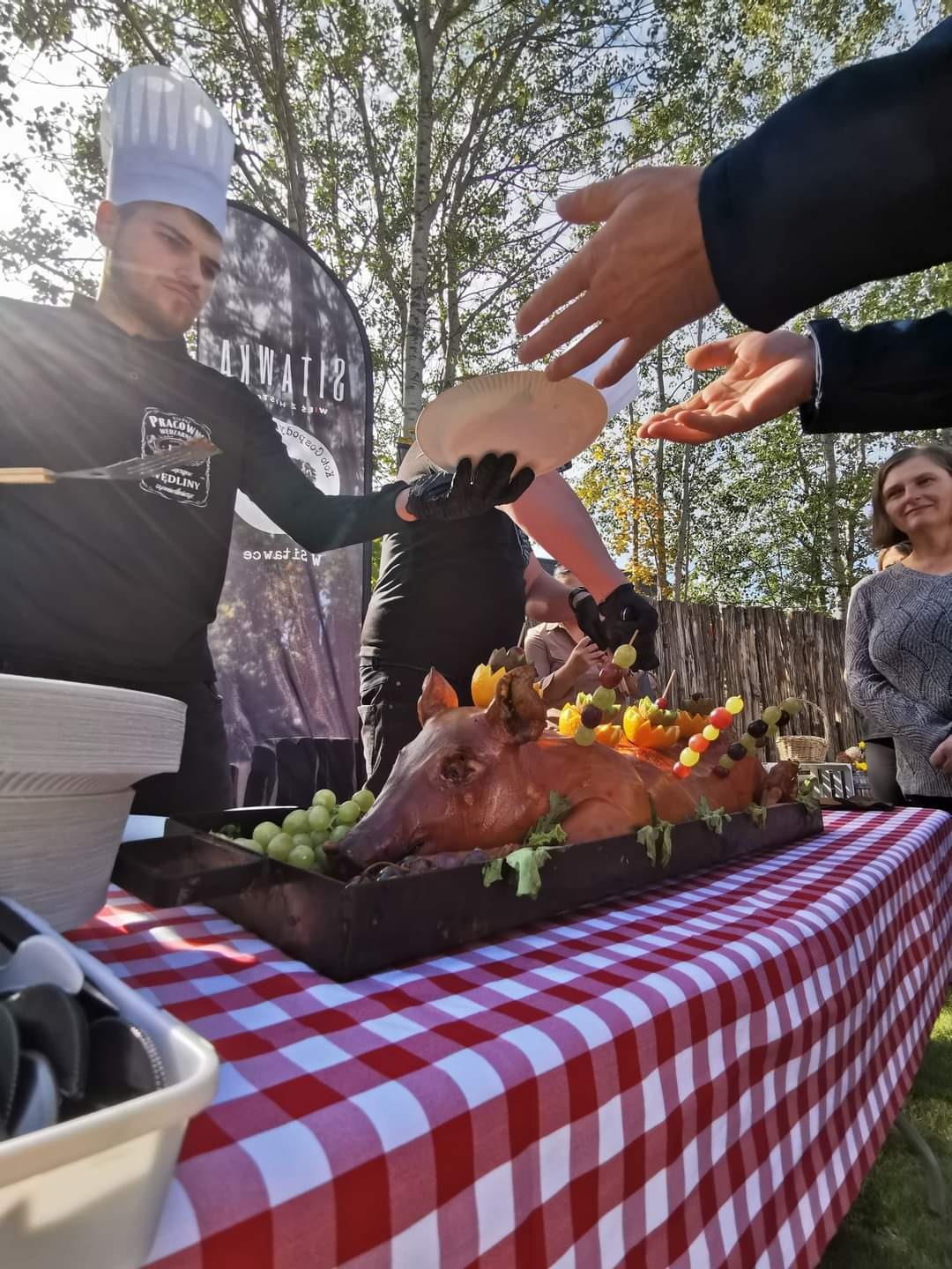Catering w Sokółce: Prosiak pieczony, dekorowany owocami, serwowany na czerwono-białym obrusie w kratę. Kucharz w czarnym uniformie i czapce kucharskiej podaje porcję na talerz.