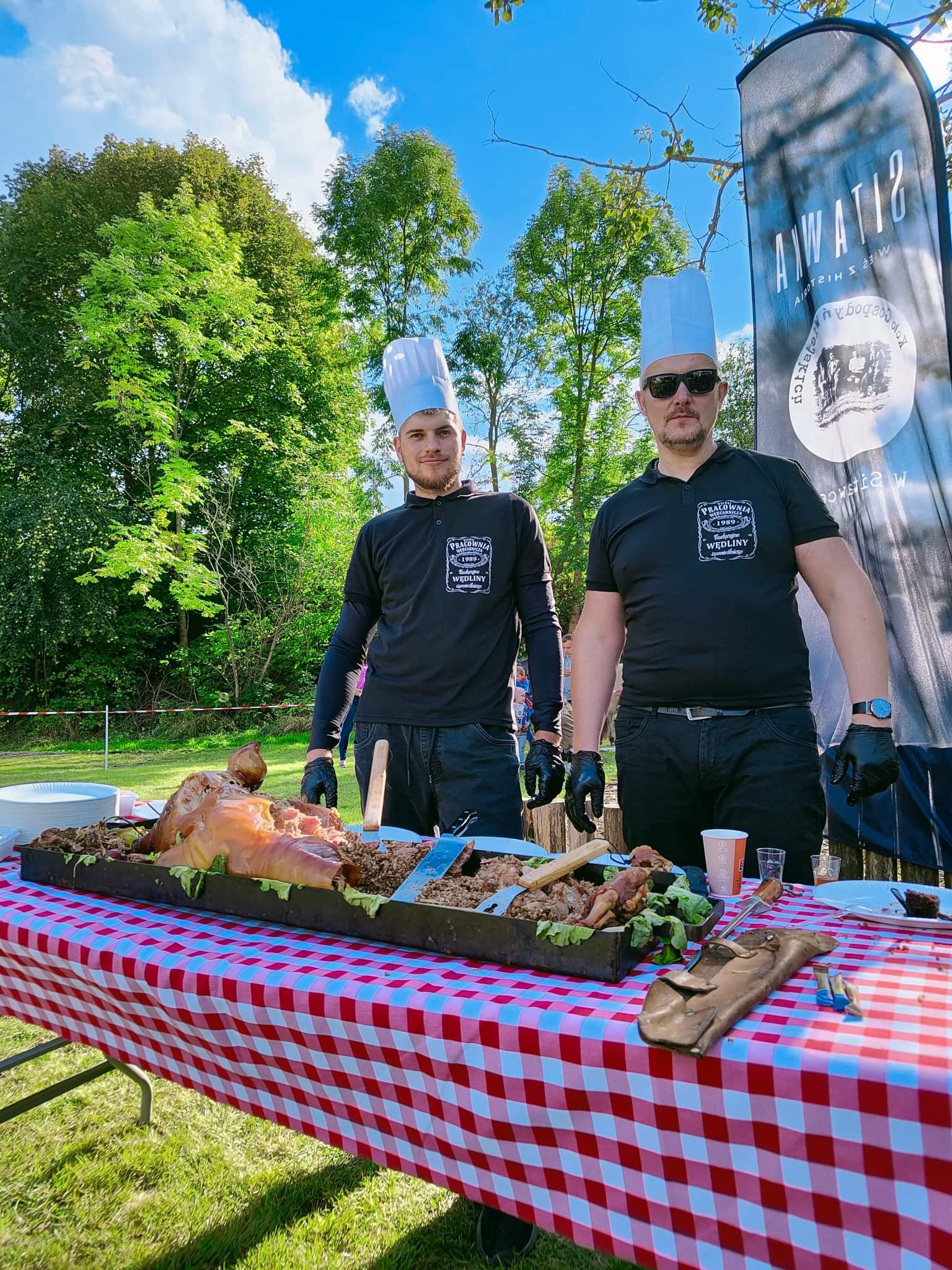 Dwóch kucharzy w czarnych uniformach i wysokich białych czapkach prezentuje pieczone prosię na metalowej tacy, ustawionej na stole z czerwono-białym obrusem w kratę, na tle zielonego trawnika i drzew.