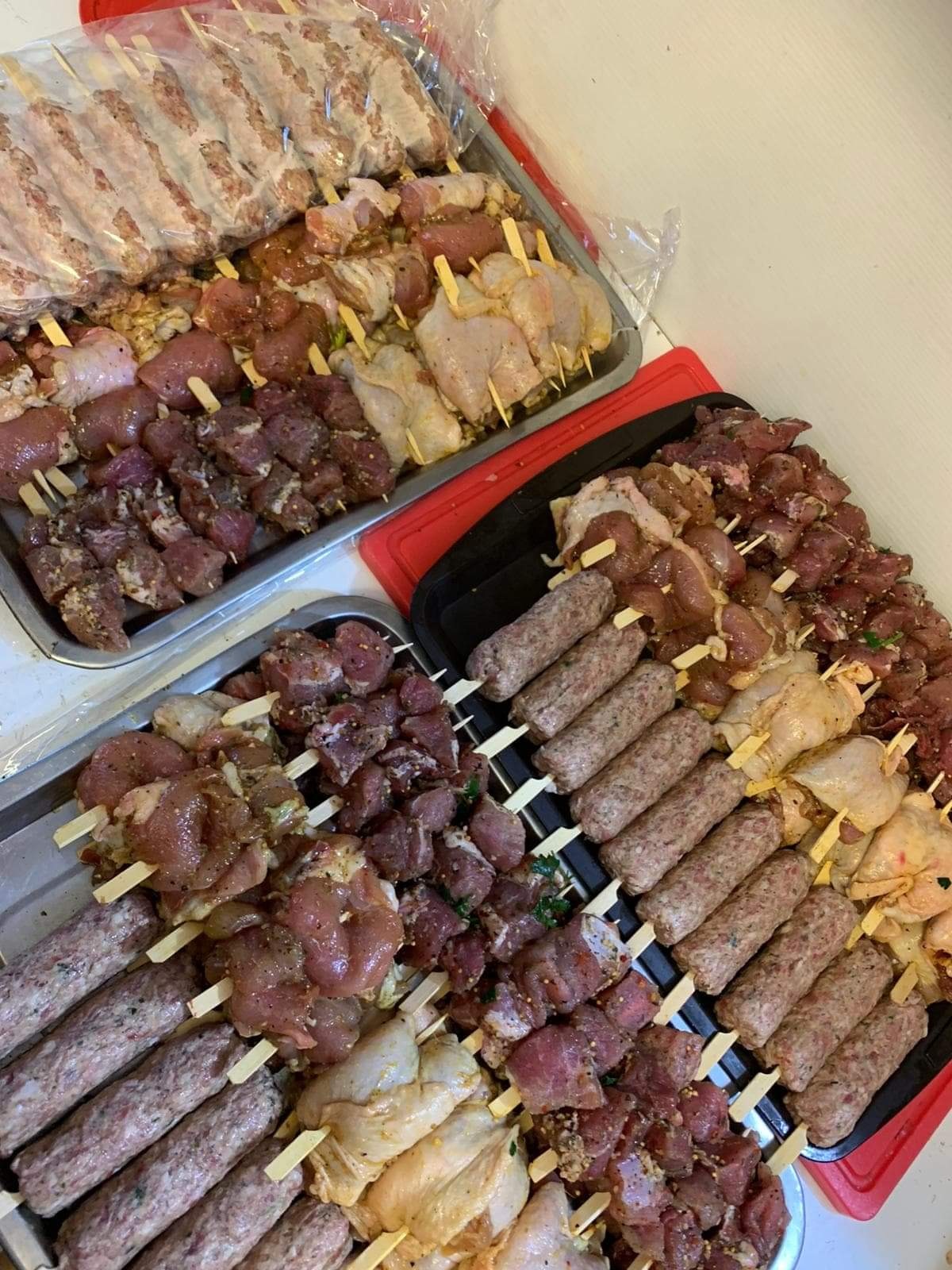 Dwa metalowe i jeden czarny pojemnik z różnymi rodzajami surowych szaszłyków i kiełbasek na drewnianych patykach, przygotowanych do grillowania lub pieczenia.