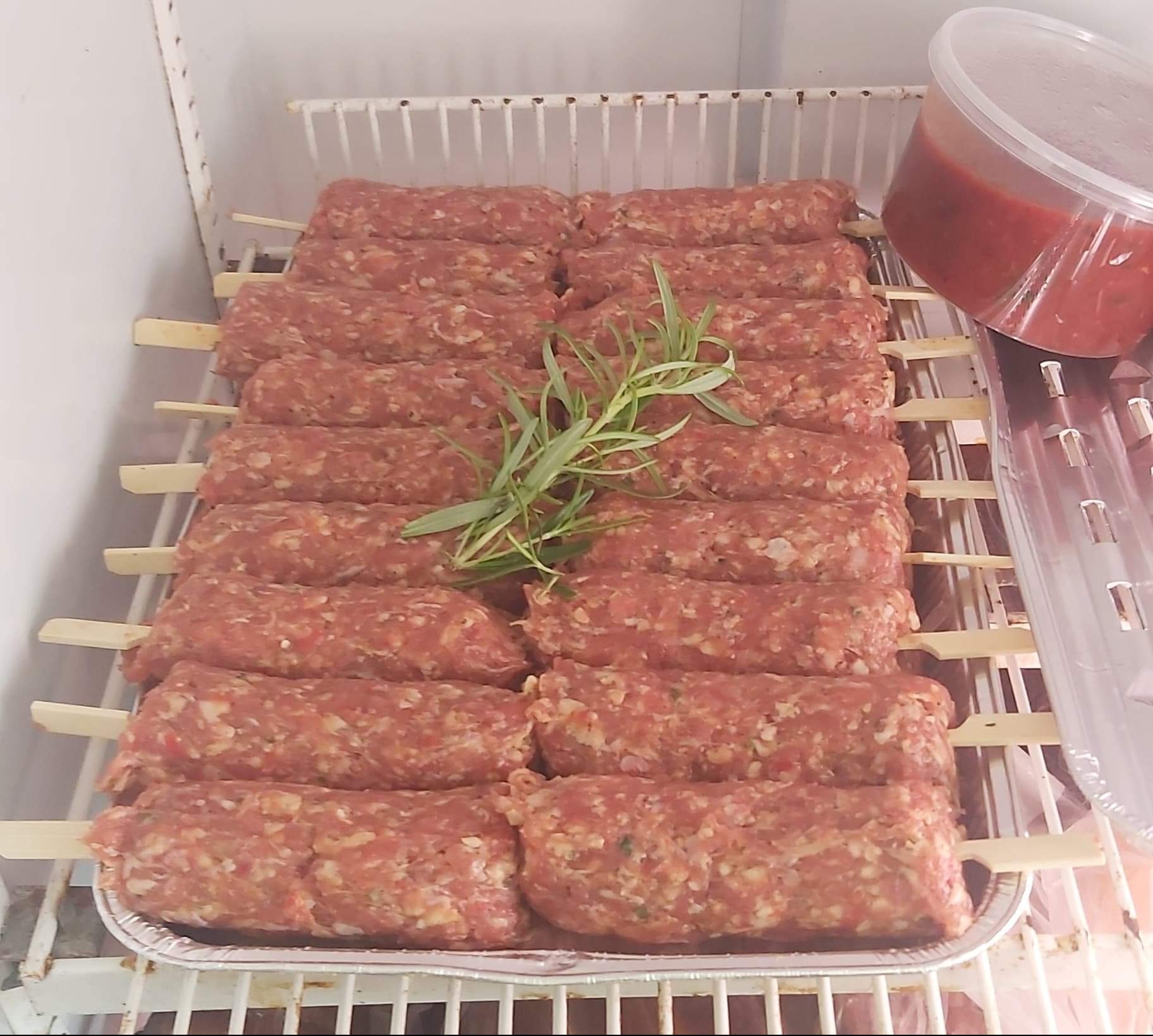 Szpikulce z surowym mięsem mielonym, udekorowane gałązką rozmarynu, ułożone na aluminiowej tacce w chłodziarce, obok pojemnik z sosem pomidorowym.