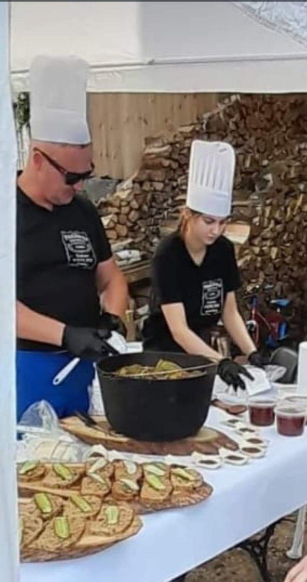 Dwoje kucharzy w czarnych koszulkach i białych czapkach przygotowuje potrawy na stole z białym obrusem. Na stole znajdują się deski z przekąskami, duży garnek z jedzeniem i małe pojemniki z sosem.