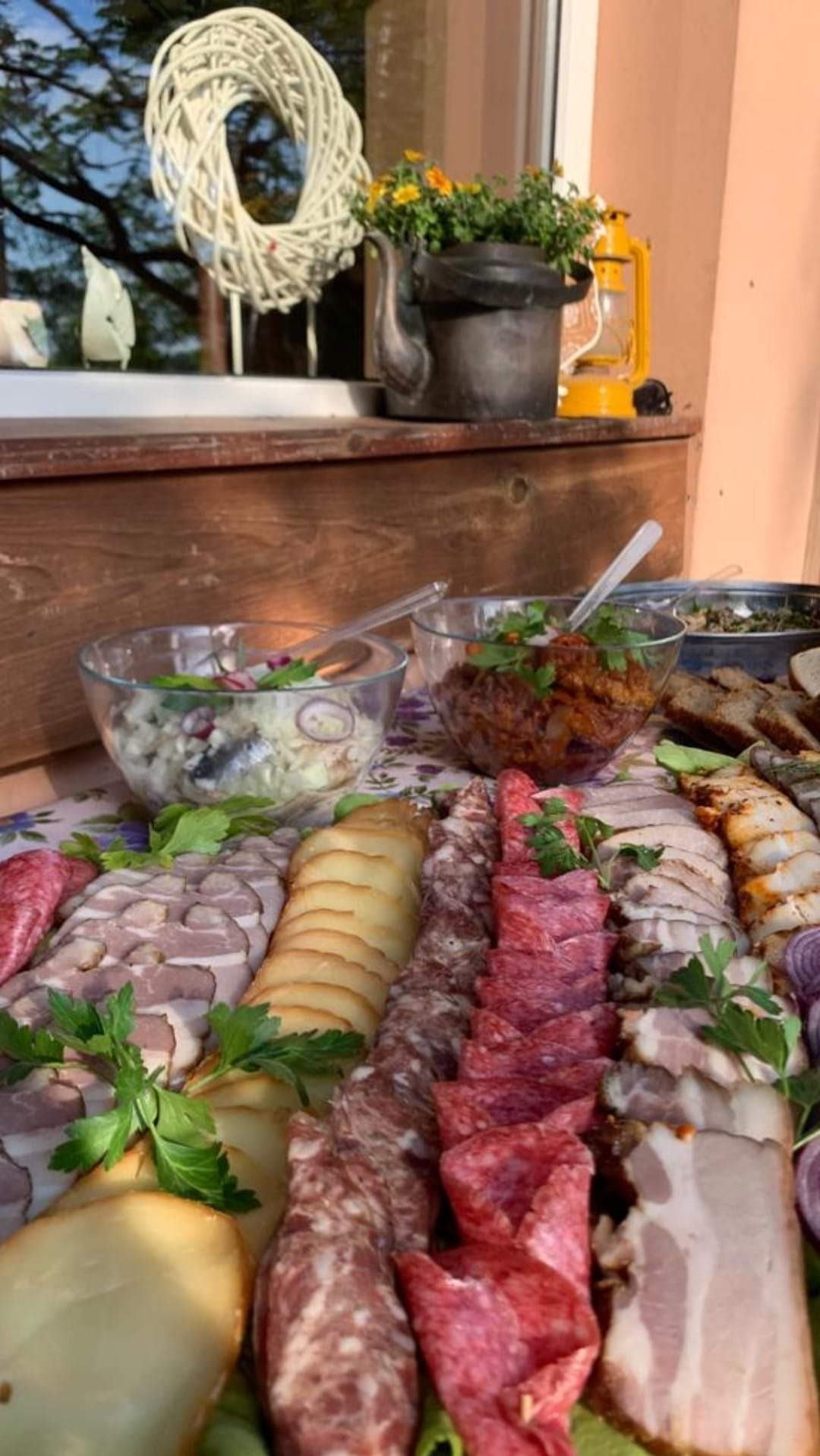 Bogato zastawiony stół z wędlinami, serami, sałatkami w szklanych miskach, dekorowany zieloną pietruszką, z widocznym wieńcem dekoracyjnym i starą lampą naftową w tle.