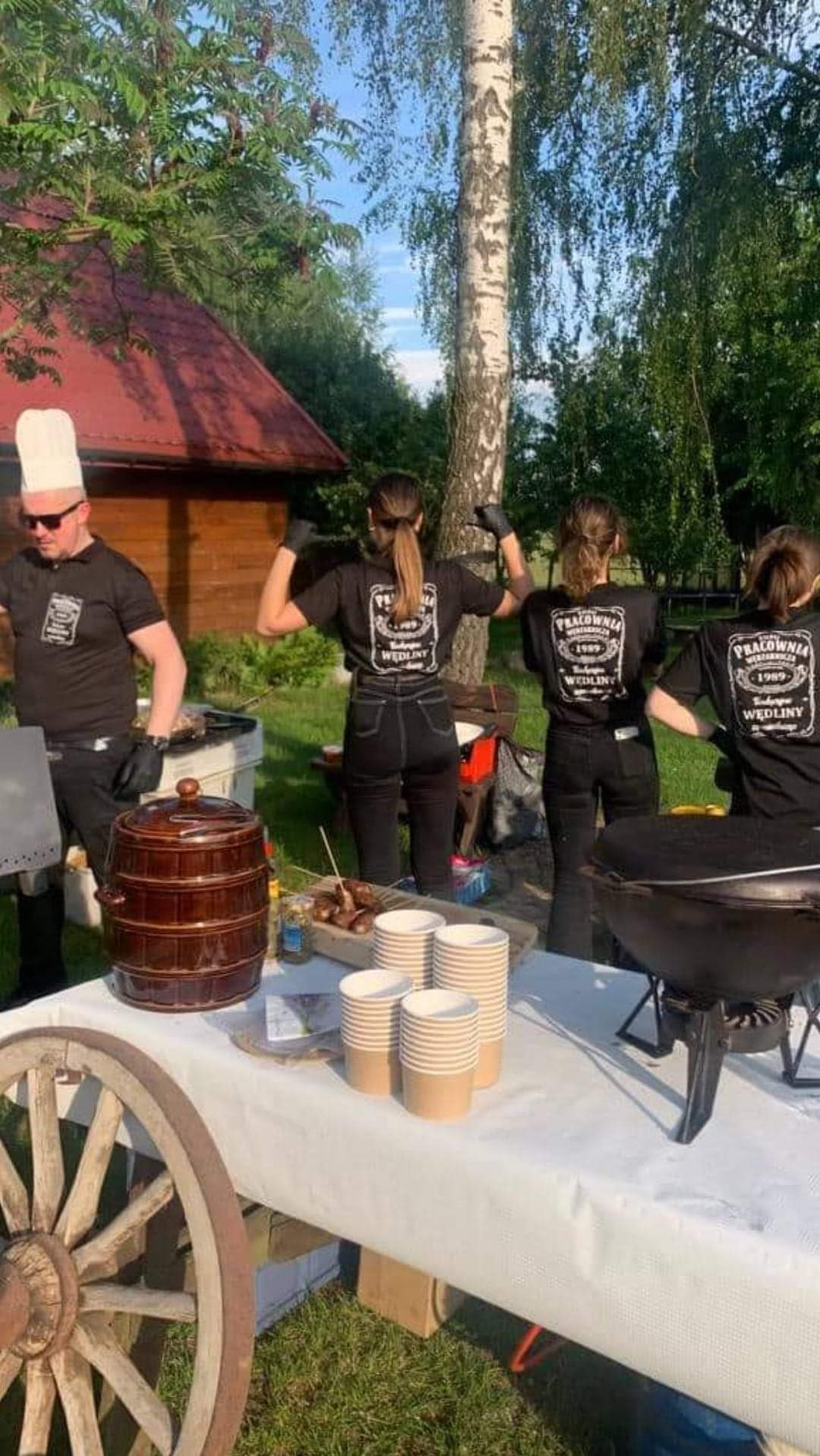 Ekipa w czarnych koszulkach z logo firmy 'Pracownia Wędlin 1989' przygotowuje jedzenie na zewnątrz, widoczny stół z przekąskami, brązowa beczka, grill węglowy i koło od wozu.