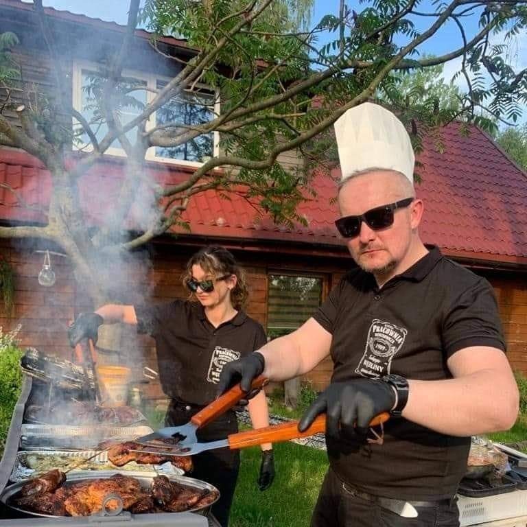 Dwóch kucharzy w czarnych koszulkach z logo firmy i okularach przeciwsłonecznych grilluje mięso na zewnątrz, unoszący się dym, w tle budynek z czerwoną dachówką.