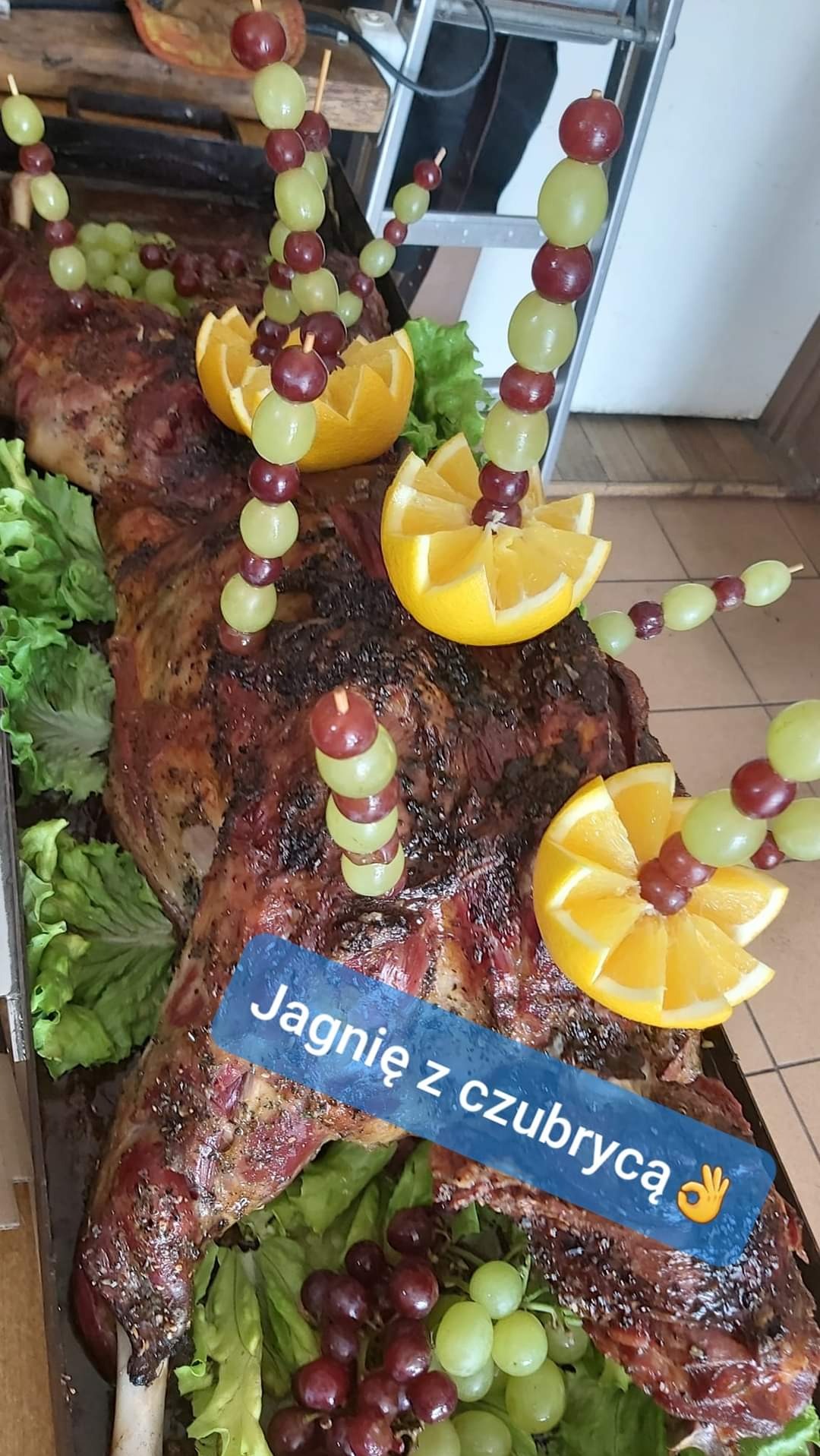 Pieczone jagnię w całości, udekorowane pomarańczami i szaszłykami z winogron, na liściach sałaty, na metalowej tacy.