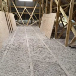 Poddasze w trakcie ocieplania, widoczne drewniane krokwie, izolacja celulozowa między legarami oraz płyty OSB przygotowane do montażu.