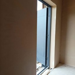 Usługi ogulno budowlane Arkadiusz Filipiak - Surowe ściany z nałożonym tynkiem w odcieniach beżu, z widocznym fragmentem szarej, segmentowej bramy garażowej w trakcie montażu, widoczne niedoskonałości posadzki.