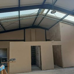 Usługi ogulno budowlane Arkadiusz Filipiak - Wnętrze hali przemysłowej w trakcie prac wykończeniowych, ściany po nałożeniu tynku, widoczne stalowe elementy konstrukcji dachu i okna dachowe, oświetlenie robocze na pierwszym planie.