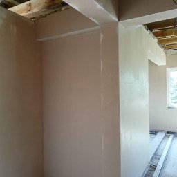 Usługi ogulno budowlane Arkadiusz Filipiak - Świeżo położona, jasna gładź na ścianach i suficie w trakcie prac wykończeniowych. Widoczne drewniane elementy konstrukcyjne sufitu i listwy przypodłogowe zabezpieczające podłogę.