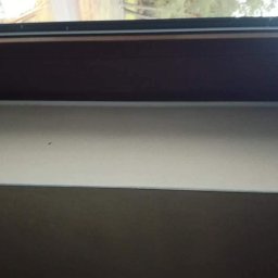 Usługi ogulno budowlane Arkadiusz Filipiak - Fragment ściany z widoczną warstwą świeżo nałożonej, jasnej masy szpachlowej, górna część ściany pomalowana na ciemny kolor, widoczne okno w tle.