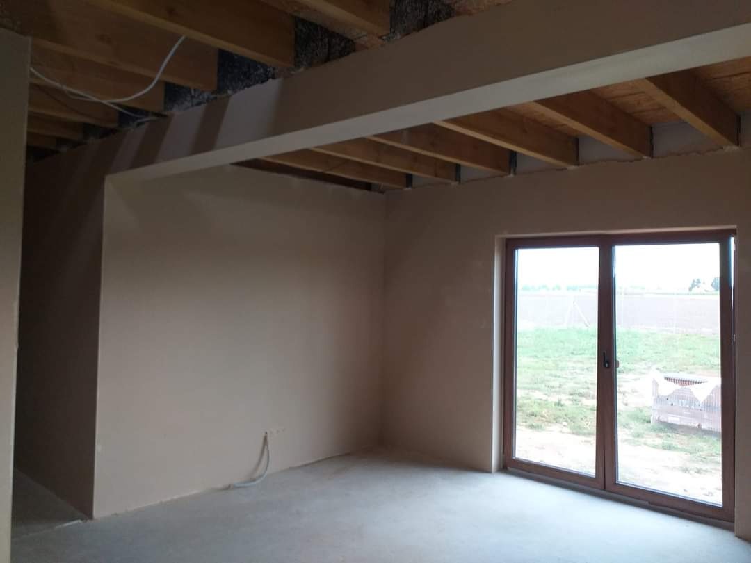 Pokój po nałożeniu tynków maszynowych, widoczne surowe drewniane belki stropowe i okno z widokiem na pole.