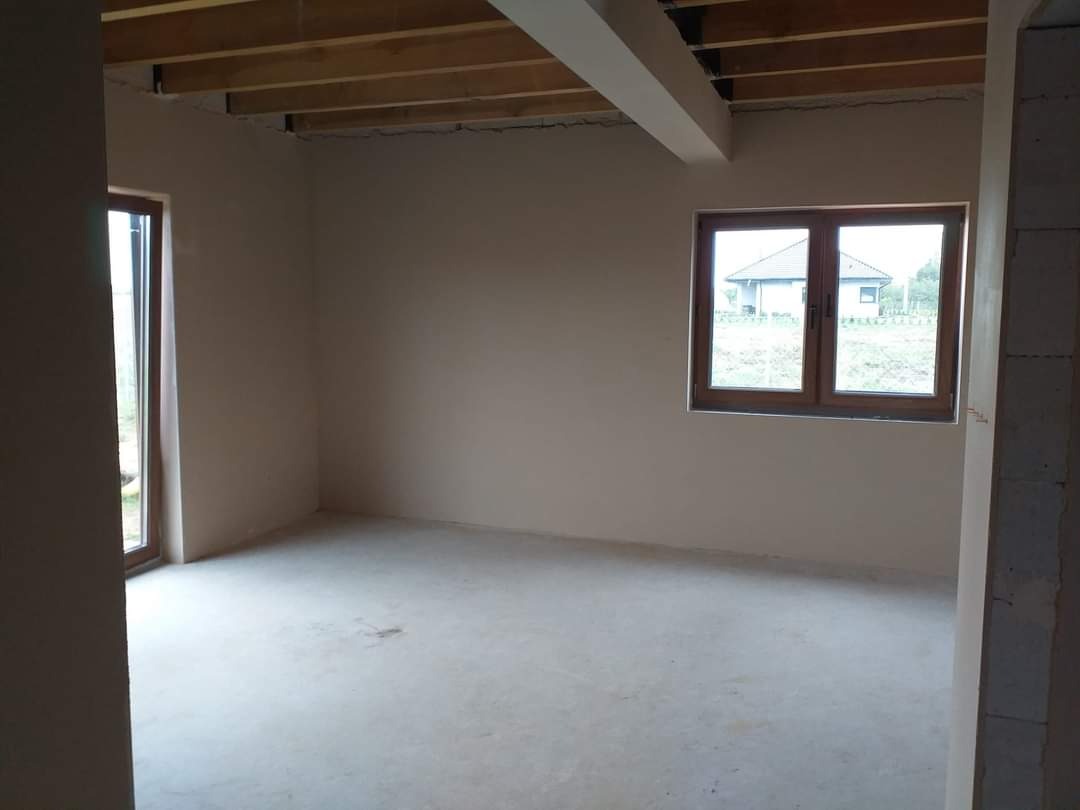 Surowe wnętrze pokoju z drewnianą konstrukcją stropu, świeżo otynkowanymi ścianami i oknem z widokiem na dom.