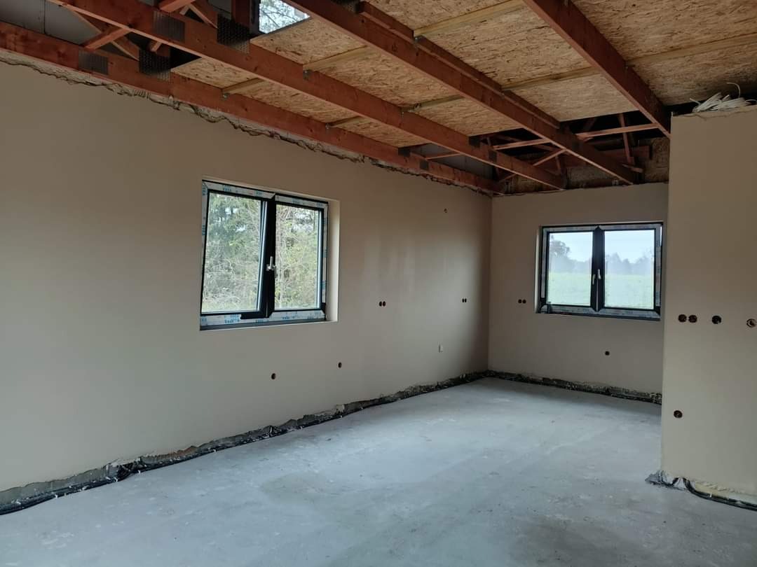 Surowe wnętrze pokoju z dwoma oknami, ściany pokryte jasnym tynkiem, widoczne instalacje elektryczne w ścianach, sufit z drewnianą konstrukcją i płytami OSB, podłoga wylewka betonowa.