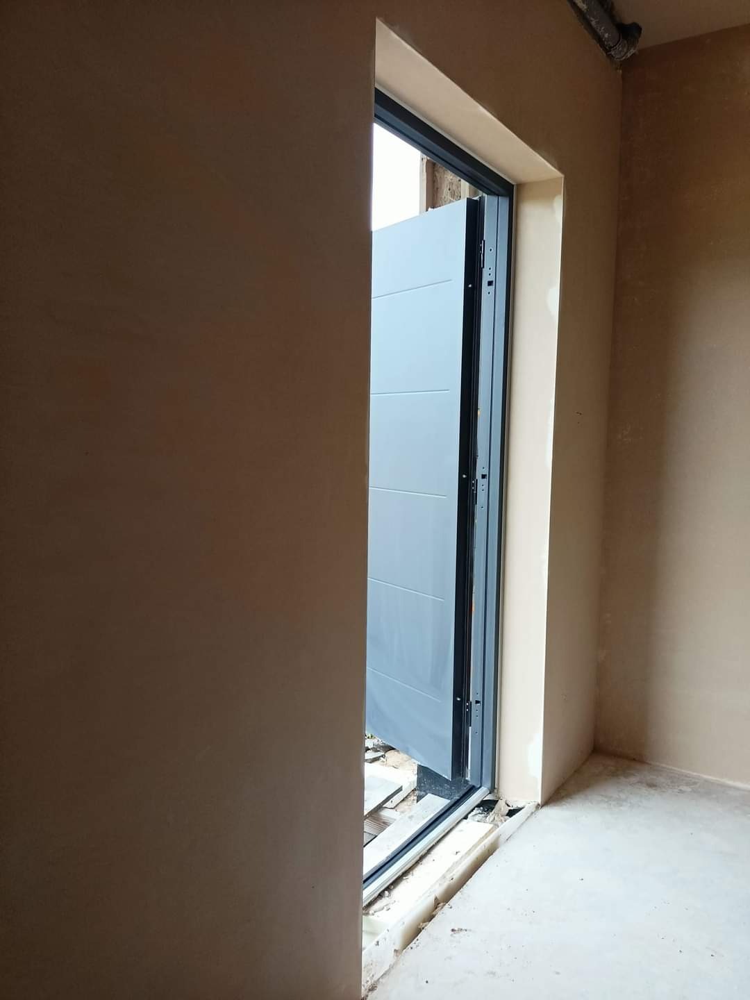 Surowe ściany z nałożonym tynkiem w odcieniach beżu, z widocznym fragmentem szarej, segmentowej bramy garażowej w trakcie montażu, widoczne niedoskonałości posadzki.