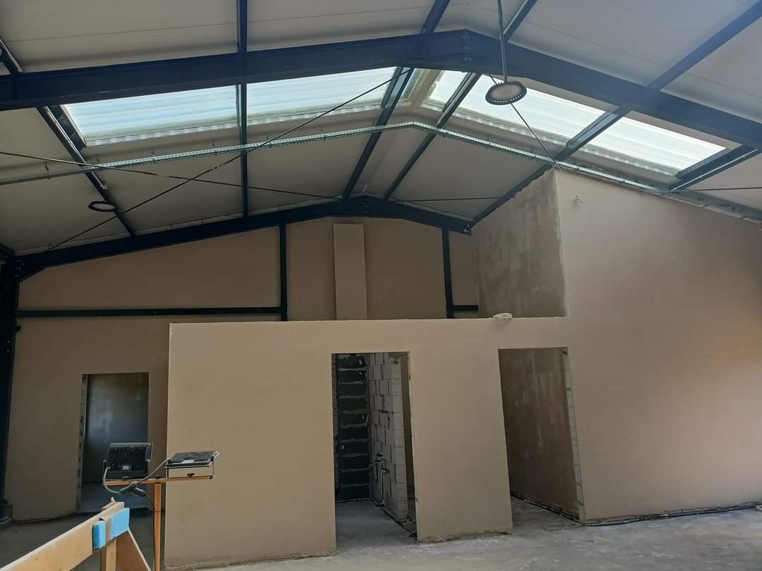 Wnętrze hali przemysłowej w trakcie prac wykończeniowych, ściany po nałożeniu tynku, widoczne stalowe elementy konstrukcji dachu i okna dachowe, oświetlenie robocze na pierwszym planie.