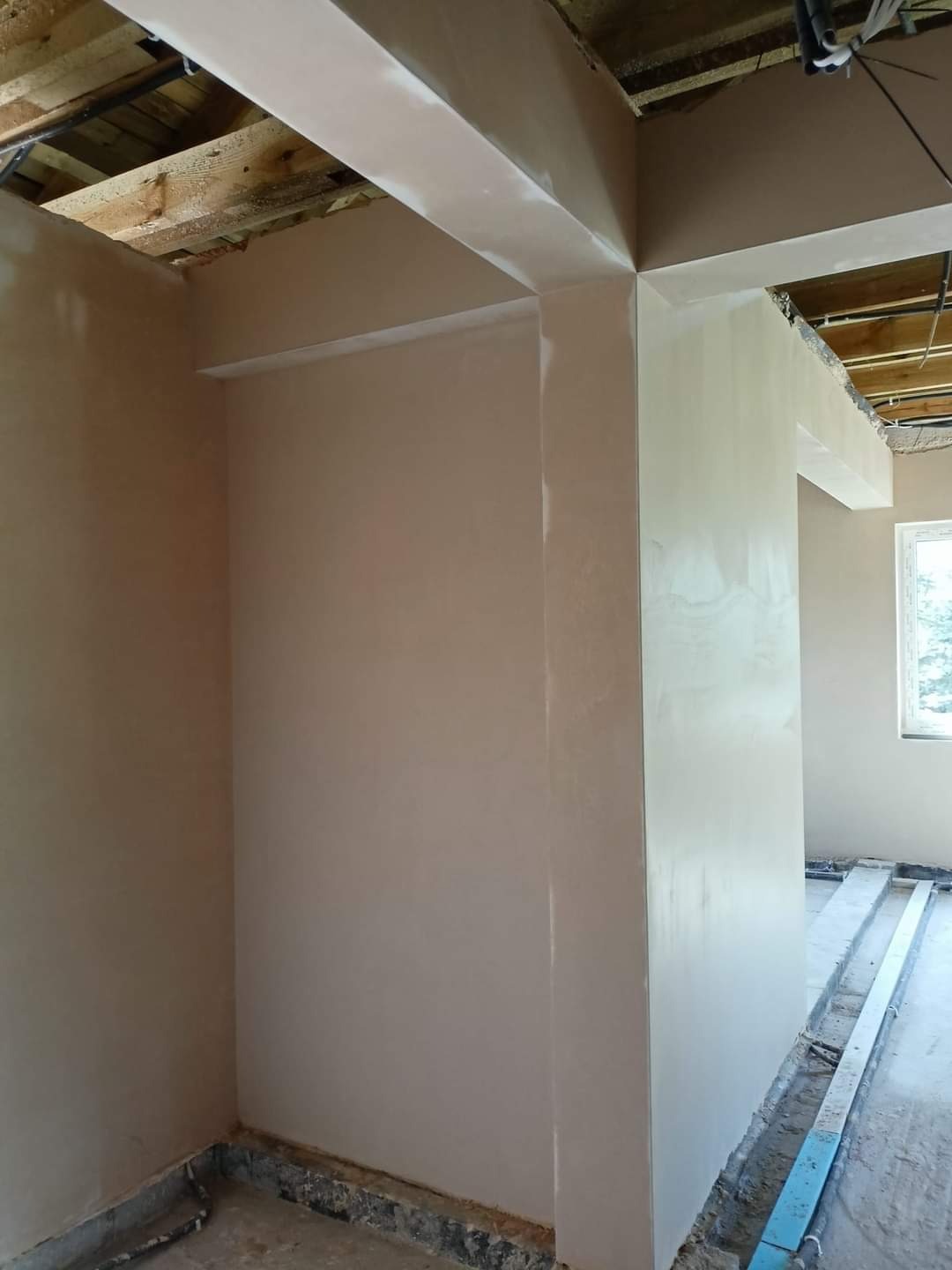 Świeżo położona, jasna gładź na ścianach i suficie w trakcie prac wykończeniowych. Widoczne drewniane elementy konstrukcyjne sufitu i listwy przypodłogowe zabezpieczające podłogę.