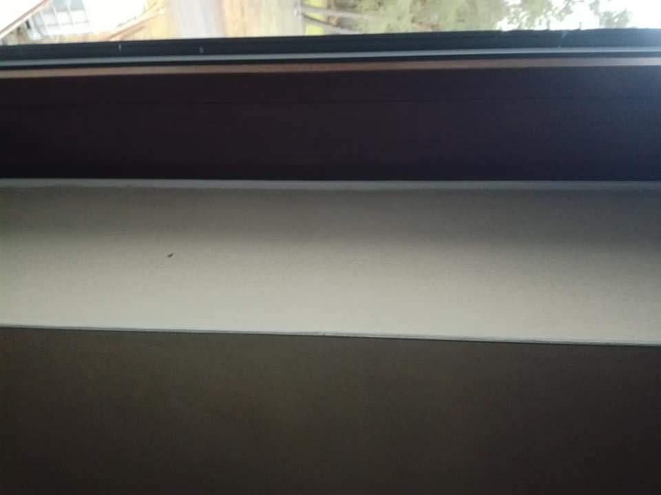 Fragment ściany z widoczną warstwą świeżo nałożonej, jasnej masy szpachlowej, górna część ściany pomalowana na ciemny kolor, widoczne okno w tle.