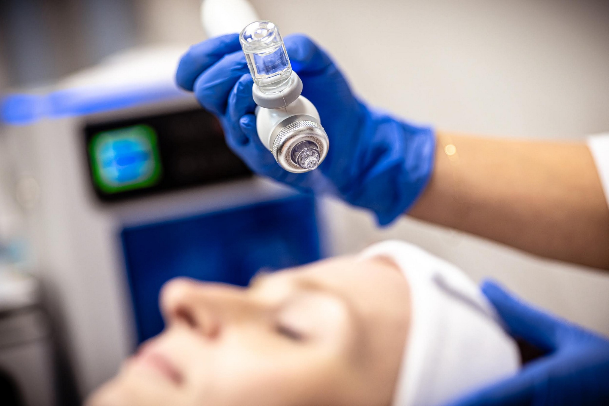 Hydrabrazja: kosmetolog w niebieskich rękawiczkach aplikuje serum na twarz klientki za pomocą urządzenia do hydrabrazji.