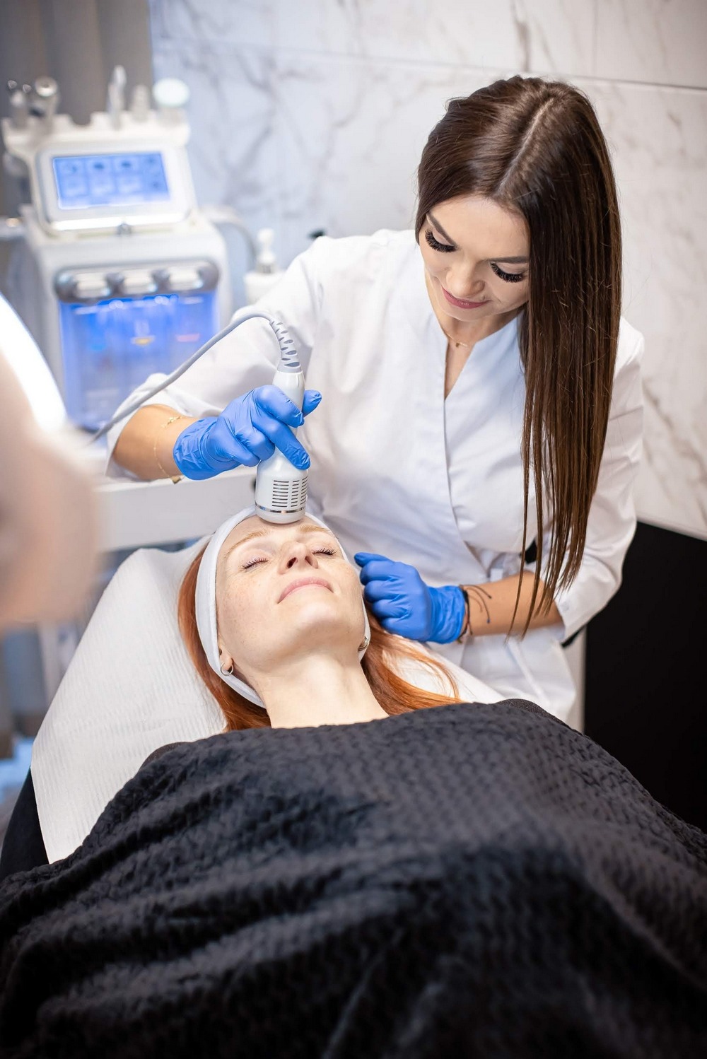 Zabieg kosmetyczny na twarzy klientki z użyciem specjalistycznego urządzenia, wykonywany przez kosmetologa w niebieskich rękawiczkach.
