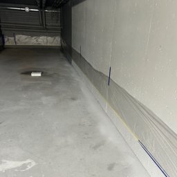 CARLO EXPERT KAROL KRZYŻANOWSKI - Przygotowane do malowania ściany w szarym pomieszczeniu, zabezpieczone folią i taśmą malarską. Widoczne instalacje pod sufitem.