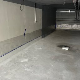 CARLO EXPERT KAROL KRZYŻANOWSKI - Przygotowane pomieszczenie do malowania: ściany zabezpieczone folią, widoczne rury pod sufitem, szpachlowana podłoga i narzędzia malarskie.