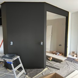 CARLO EXPERT KAROL KRZYŻANOWSKI - Ciemnoszara ściana w trakcie malowania z widoczną drabiną, kuwetą malarską i zabezpieczoną folią podłogą. Żółta taśma malarska na krawędziach. Świeżo pomalowane.