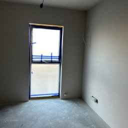 CARLO EXPERT KAROL KRZYŻANOWSKI - Pokój po gładziowaniu ścian, okno zabezpieczone folią i taśmą malarską. Ściany w jasnym odcieniu szarości, surowa podłoga. Widoczne przewody.