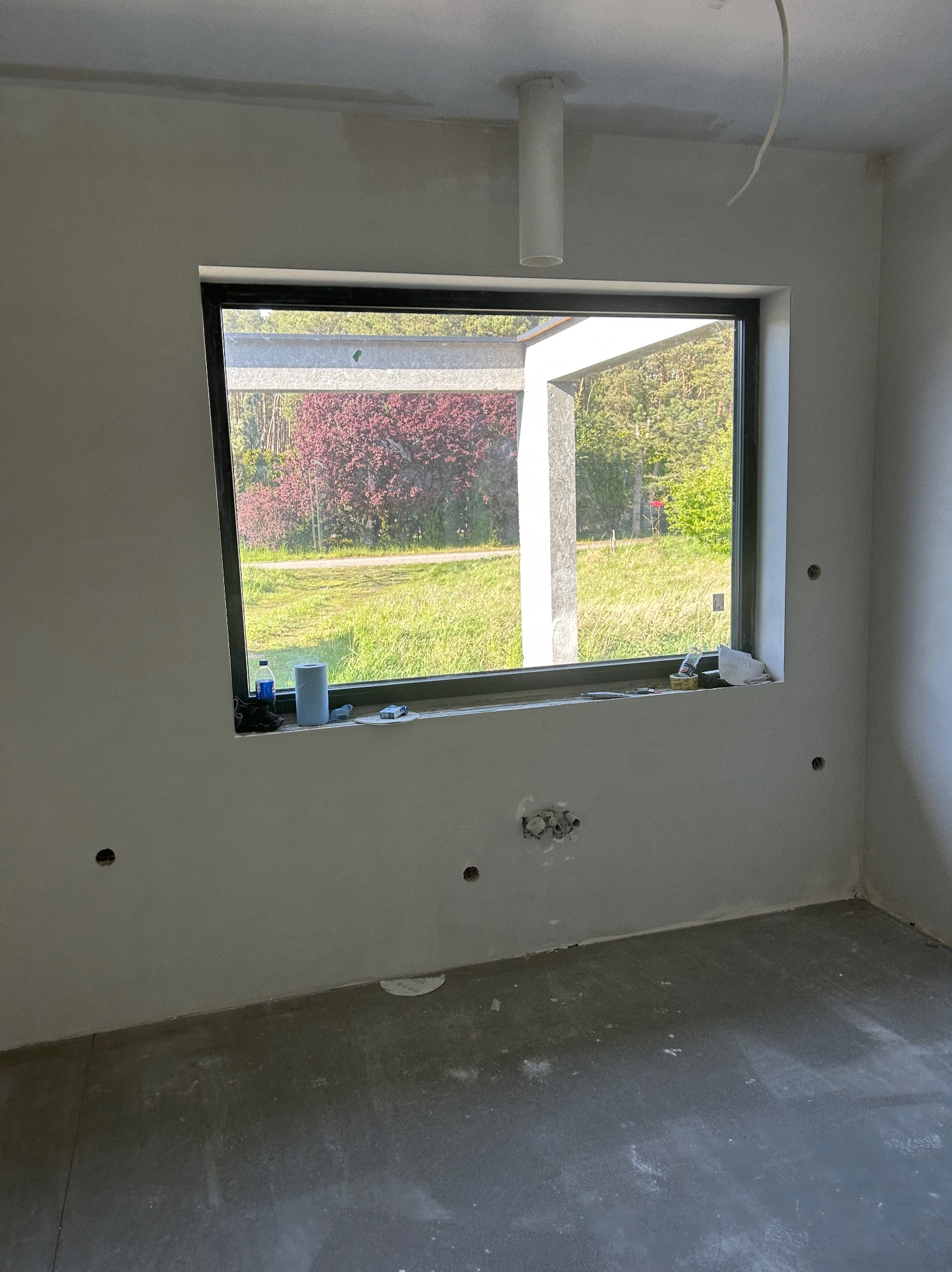 Wnętrze w trakcie prac wykończeniowych: ściany po gładzi, widoczne puszki elektryczne, okno z widokiem na zieleń. Podłoga surowa, przygotowana do dalszych prac.