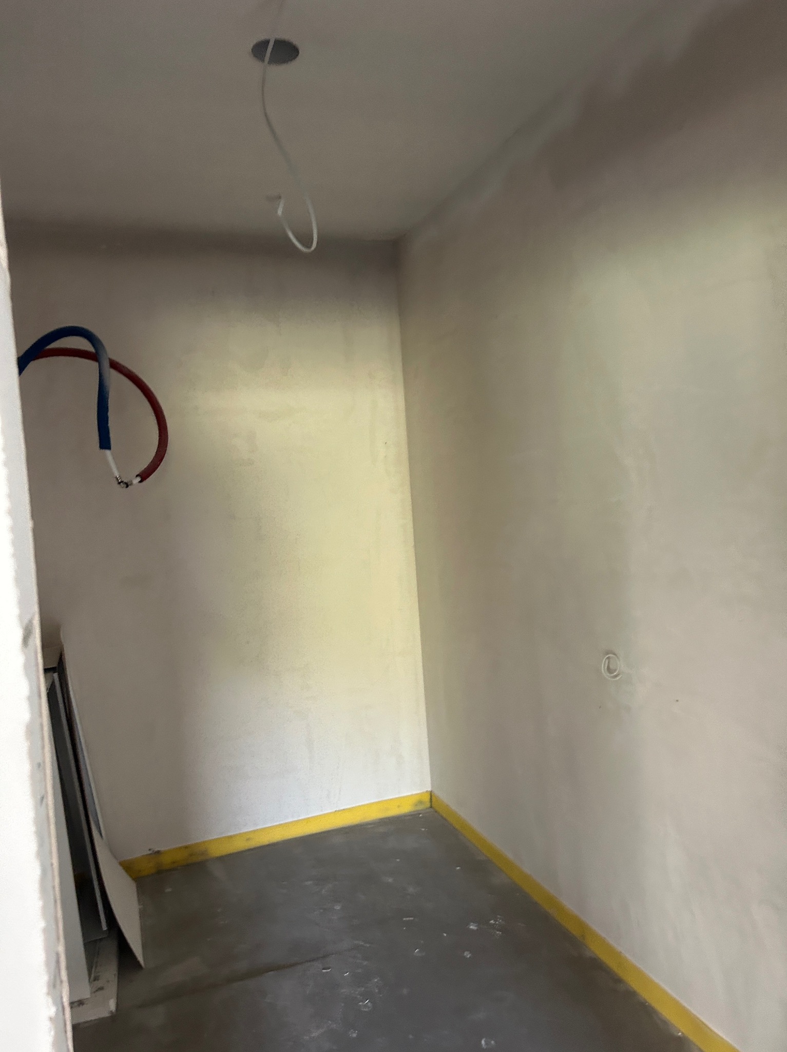 Wnętrze pokoju po nałożeniu gładzi, widoczne kable i rury. Podłoga zabezpieczona żółtą taśmą. Ściany gładkie, przygotowane do malowania.